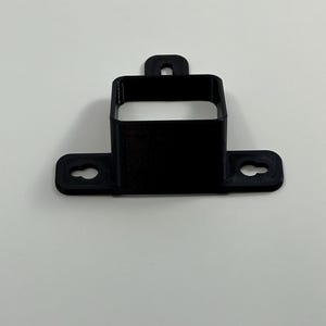 May include: A black, 3D-printed bracket with a rectangular opening and a mounting tab. The bracket has two circular holes on the base for screws. The overall design is simple and functional.
