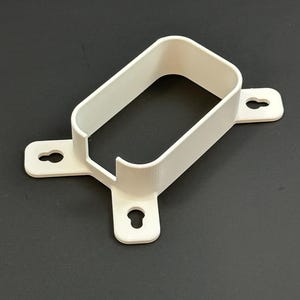 Puede incluir: Un soporte rectangular blanco con esquinas redondeadas y tres orificios de montaje en forma de ojo de cerradura. El soporte tiene una abertura en forma de U en el centro y está hecho de un material sólido y opaco. El objeto está sobre un fondo oscuro.