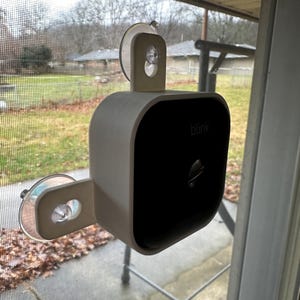May include: A white and black Blink security camera with a suction cup mount attached to a window. The camera has the word "blink" printed on the front. The background shows a residential area with trees and a cloudy sky.