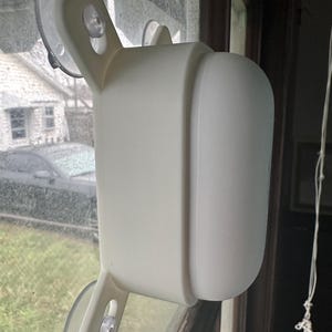 Puede incluir: Dispositivo de plástico blanco con dos ventosas a cada lado, montado en una ventana. El dispositivo tiene una superficie redondeada y lisa.