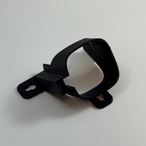 Może przedstawiać: Czarny, drukowany w 3D plastikowy wspornik o zakrzywionym kształcie. Posiada dwa otwory montażowe i prostokątny odcinek. Wspornik jest przeznaczony do dopasowania wokół obiektu, z gładkim, matowym wykończeniem.
