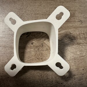 Puede incluir: Soporte de pared de plástico blanco con cuatro pestañas y cuatro agujeros. El soporte está diseñado para sujetar un objeto cuadrado.