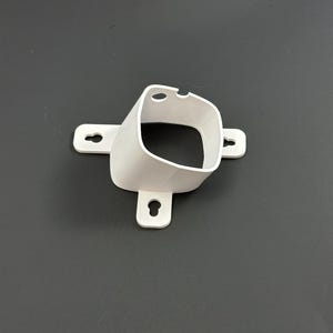 Op de afbeelding: Een witte, 3D-geprinte kunststof beugel met een vierkante opening en drie montage-lipjes. De beugel heeft een gebogen ontwerp en wordt waarschijnlijk gebruikt voor het monteren of vastzetten van een object. De achtergrond is donkergrijs.