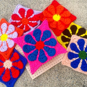 May include: Eight crocheted flower squares in various colors, including purple, pink, blue, red, orange, and brown. The squares are arranged on a wooden surface.