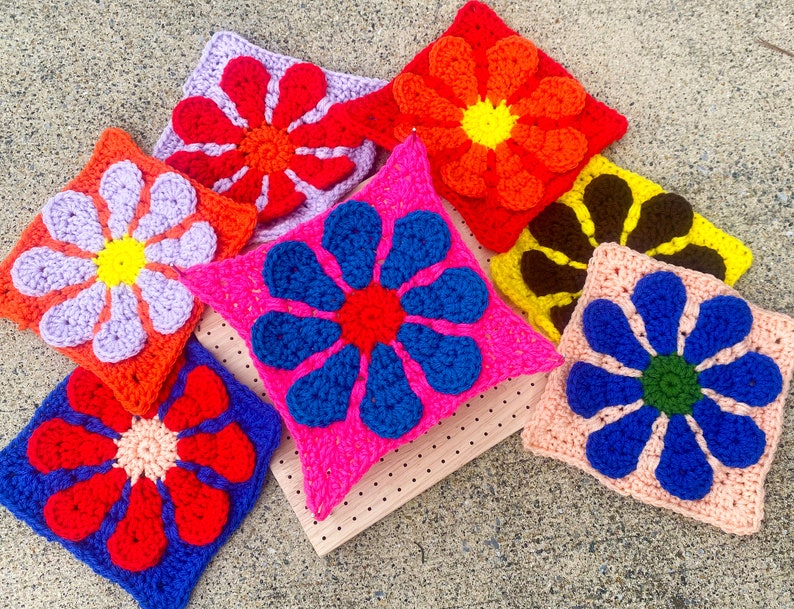 May include: Eight crocheted flower squares in various colors, including purple, pink, blue, red, orange, and brown. The squares are arranged on a wooden surface.