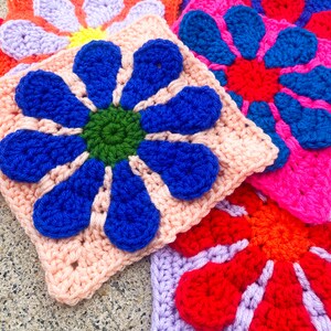 May include: Four crocheted flower squares in different colors: pink with blue flower, purple with red flower, pink with green flower, and red with orange flower.