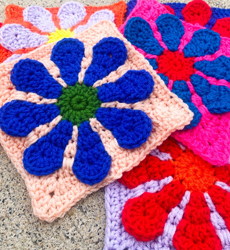 May include: Four crocheted flower squares in different colors: pink with blue flower, purple with red flower, pink with green flower, and red with orange flower.