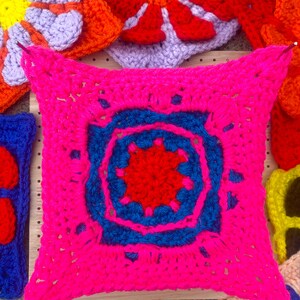 May include: A bright pink crocheted square with a blue and red center design. The square is surrounded by other crocheted squares in various colors, including purple, orange, and yellow.