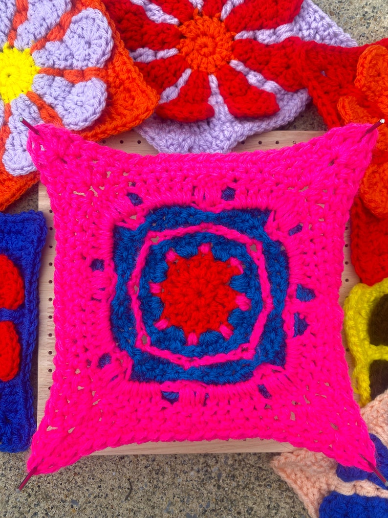 May include: A bright pink crocheted square with a blue and red center design. The square is surrounded by other crocheted squares in various colors, including purple, orange, and yellow.