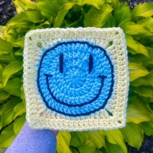 Peut inclure: Un carré crocheté avec un smiley bleu sur fond jaune. Le carré est tenu par une main.