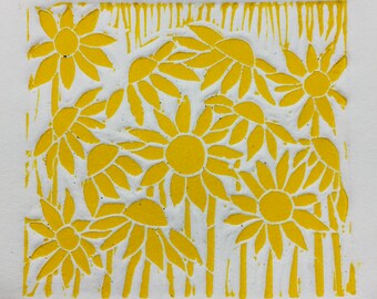 May include: A yellow linoleum block print of a field of sunflowers. The print features multiple sunflowers with yellow petals and centers. The background is a white paper with a striped pattern.