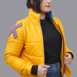 May include: A yellow puffer jacket with a blue interior and a purple "X" patch on the shoulder. The jacket has a black ribbed cuff and a blue zipper. The model is wearing a black shirt and blue jeans.