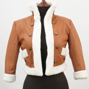 Könnte beinhalten: Eine braune Kunstlederjacke mit einem weißen Kunstfellkragen und Futter. Die Jacke hat eine verkürzte Länge und verfügt über Knopfverschlüsse.