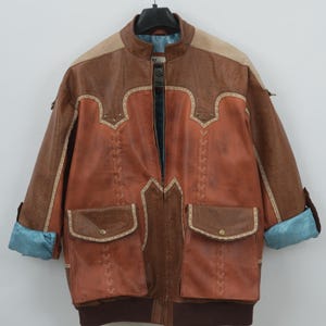 May include: A vintage brown leather jacket with a unique design. The jacket features a mix of textures and shades of brown, with decorative stitching and two front pockets. The cuffs are rolled up, revealing a blue lining. The jacket is hanging on a black hanger.