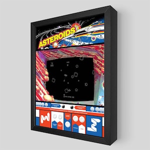 Puede incluir: Impresión enmarcada del clásico juego de arcade Asteroids. La obra de arte presenta el logotipo del juego, una nave espacial y una pantalla negra que muestra los gráficos del juego. El marco es negro y la impresión tiene una estética retro.