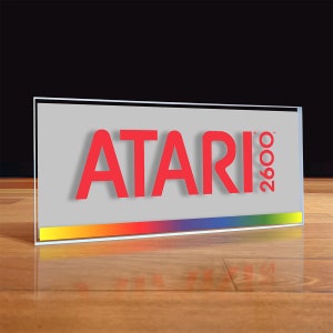 May include: A clear glass plaque with a red "ATARI 2600" logo. The logo has a slight shadow effect. A rainbow stripe is at the bottom of the plaque.