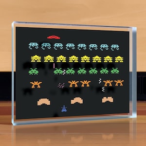 May include: A black and white pixelated image of a classic arcade game, Space Invaders, in a glass frame. The image shows the game in progress with the player's ship at the bottom of the screen and the invading aliens moving down from the top. The aliens are in different colors, including green, yellow, and blue.