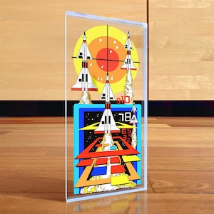 Puede incluir: Un bloque acrílico colorido con un diseño retro de temática espacial. La obra de arte incluye cohetes despegando de una plataforma, un objetivo y el número "18". La paleta de colores incluye rojo, naranja, amarillo, azul y blanco.