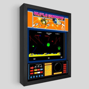 Puede incluir: Obra de arte enmarcada con el clásico juego de arcade "Missile Command". La obra muestra el título del juego en naranja y blanco, junto con el logotipo de Atari. La pantalla del juego tiene un fondo negro con gráficos verdes y rojos. El marco es negro.