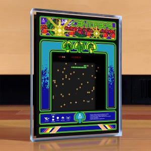 May include: A framed Centipede arcade game cabinet. The cabinet features a black screen displaying the game's graphics, with a blue and green border. The top of the cabinet has the word "Centipede" in green, and the Atari logo is visible.