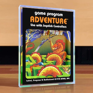 Puede incluir: Un cartucho de juego Atari vintage para Adventure, con una ilustración colorida de un dragón y un laberinto. El texto en el cartucho dice "game program ADVENTURE Use with Joystick Controllers Label, Program & Audiovisual ©1978 ATARI, INC."