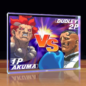 May include: A video game screen showing a versus match between two characters. The character on the left is Akuma, a red-haired fighter with a fierce expression. The character on the right is Dudley, a boxer with a mustache and a blue boxing glove. The text "VS" is displayed in the center of the screen, with "1P Akuma" and "Dudley 2P" displayed at the bottom of the screen.