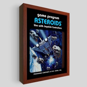 Puede incluir: Una imagen enmarcada del clásico videojuego Atari Asteroids. La imagen muestra una nave espacial volando por el espacio, esquivando asteroides. El texto en el marco dice "game program ASTEROIDS Use with Joystick Controllers PROGRAM CONTENTS ©1981 ATARI, INC."