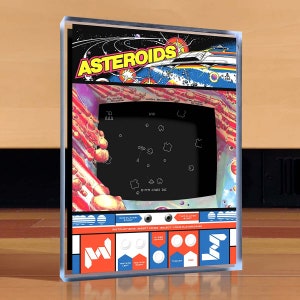 Puede incluir: Un gabinete de juego de arcade de estilo retro con un diseño azul y rojo. El gabinete presenta una pantalla que muestra el juego clásico "Asteroids" con una nave espacial y asteroides. El gabinete tiene botones para "Inicio de un jugador", "Inicio de dos jugadores", "Hiperespacio", "Girar a la izquierda", "Girar a la derecha", "Empuje", y "Fuego".