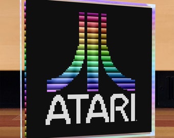 Puede incluir: Un logotipo de Atari en píxeles blanco y negro con un joystick de Atari de arcoíris en el centro. El logotipo dice "ATARI".