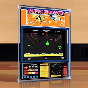 Puede incluir: Un juego de arcade enmarcado con el clásico juego "Missile Command". La sección superior muestra el título y los gráficos del juego, mientras que el centro muestra la pantalla de juego con una puntuación alta de 7500. La sección inferior incluye controles de lanzamiento e instrucciones.