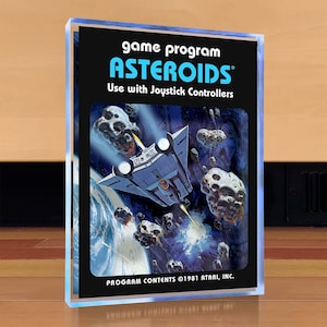 Puede incluir: Un cartucho de juego Atari vintage para Asteroids. El cartucho es negro con una etiqueta azul y blanca. La etiqueta muestra una nave espacial volando a través de un campo de asteroides. El texto en la etiqueta dice "game program ASTEROIDS Use with Joystick Controllers PROGRAM CONTENTS ©1981 ATARI, INC."