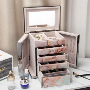 May include: A jewellery box with a mirrored lid and drawers. The drawers have a pink and gold marble design. The box is open, revealing jewellery inside, including necklaces, bracelets, and rings. The box is on a white surface.