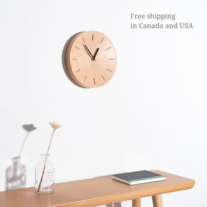 May include: A light wood wall clock with black hands and a simple design. The clock is hanging on a white wall above a wooden table with a vase and a book.