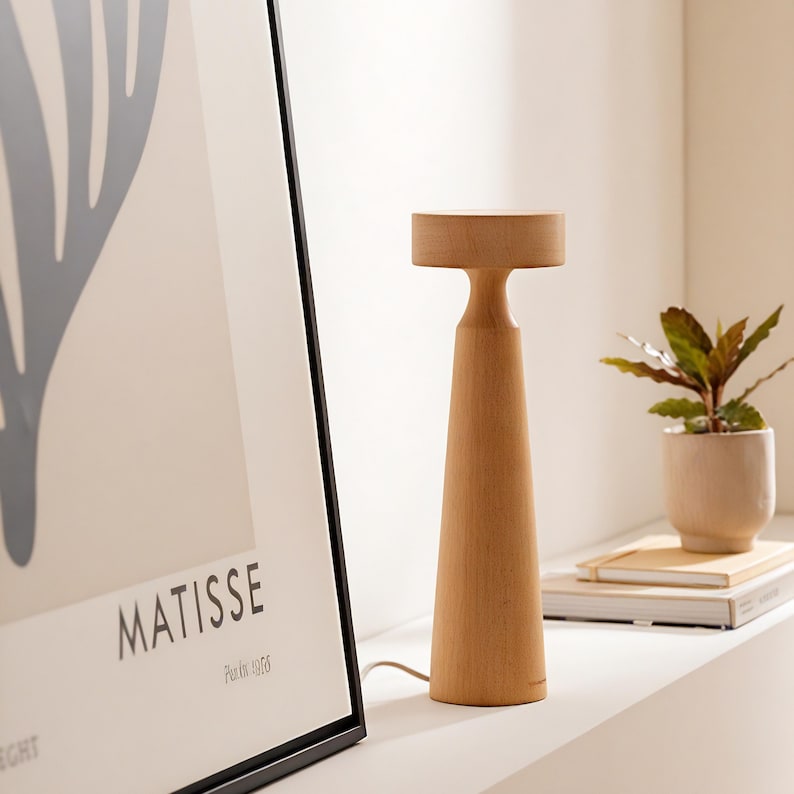 K&ouml;nnte beinhalten: Eine Tischlampe aus Holz mit einer nat&uuml;rlichen Holzoptik. Die Lampe hat einen runden Sockel und einen zylindrischen Schirm. Die Lampe steht auf einer wei&szlig;en Oberfl&auml;che neben einem gerahmten Druck mit dem Text "MATISSE" und "Paris, 1906".