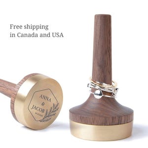 May include: Two ring holders made of dark brown wood and brushed gold metal. One holder stands upright with three silver rings on its tapered post. The other lies on its side, showing a circular gold base engraved with "ANNA & JACOB 07.27.2023".
