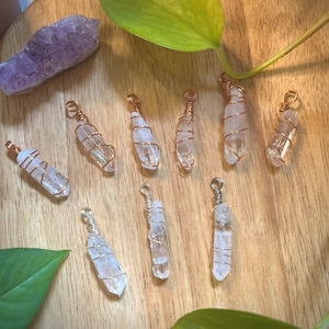 May include: Nine clear quartz crystal pendants wrapped in copper wire. The pendants are arranged on a wooden surface with a green plant in the background.