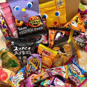 Pode incluir: Uma coleção de snacks japoneses variados numa caixa de cartão, com embalagens coloridas e gráficos temáticos de Halloween, incluindo Tohato Caramel Corn, chocolates Meiji Apollo, Pocky e vários doces embalados individualmente.