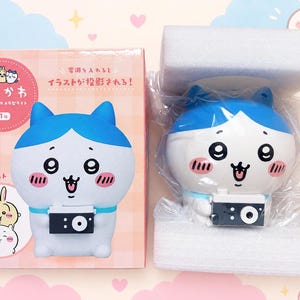 May include: A Chiikawa character light featuring a blue and white cat-like figure holding a small black camera. The product is shown inside a clear plastic bag nestled between two white foam blocks, next to its pink and white illustrated box.