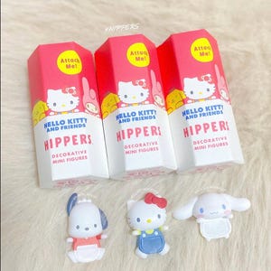 Pode incluir: Três caixas hexagonais cor-de-rosa e brancas rotuladas como Hello Kitty and Friends HIPPERS Decorative Mini Figures com autocolantes amarelos Attach Me!. Por baixo, encontram-se três pequenas figuras de plástico: um cão branco, a Hello Kitty e uma personagem branca de orelhas compridas.