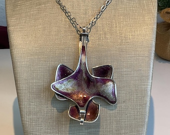 May include: A silver-toned pendant necklace with a large, abstract floral design. The pendant features a purple and silver enamel finish with a textured pattern. The necklace is displayed on a mannequin.