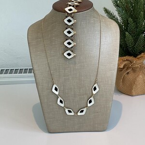 Peut inclure: Un ensemble collier et bracelet de couleur or. Les bijoux présentent des pendentifs blancs en forme de losange avec des centres noirs. Le bracelet est exposé sur un présentoir à bijoux en bois, tandis que le collier est suspendu à un mannequin.