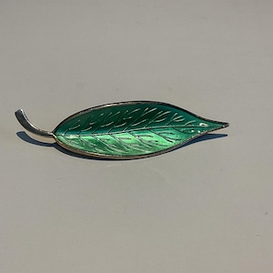 Puede incluir: Broche con forma de hoja con un vibrante acabado de esmalte verde y un borde de metal plateado. El broche tiene un diseño detallado de venas de hoja y un tallo curvo. Un accesorio inspirado en la naturaleza.