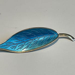 Puede incluir: Broche en forma de hoja con borde y tallo dorados. La hoja está rellena de esmalte azul degradado, con vetas azul oscuro. Un accesorio elegante para una chaqueta o bufanda.