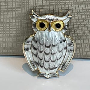 Puede incluir: Un broche de búho en metal dorado con detalles en esmalte blanco y acentos de plumas negras. El búho tiene ojos prominentes de esmalte amarillo con centros negros y un acabado texturizado en las alas y el cuerpo, creando un diseño detallado.
