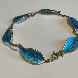 Puede incluir: Pulsera plateada con eslabones en forma de hojas con esmalte azul vibrante. Las hojas tienen vetas detalladas y están conectadas por eslabones de metal dorado. La pulsera se asegura con un cierre.