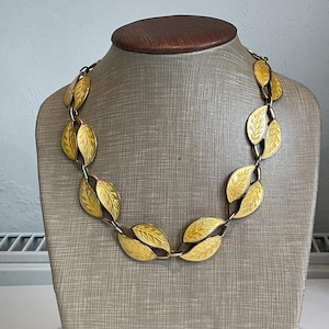 Puede incluir: Un collar dorado con colgantes en forma de hojas. Cada hoja tiene vetas detalladas y está conectada por pequeños eslabones. El collar se exhibe en un maniquí, mostrando su diseño. Las hojas tienen un tono amarillo.