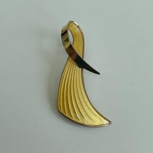Peut inclure: Broche dorée en forme de ruban. La broche présente une partie inférieure texturée en forme d'éventail et une section supérieure lisse et incurvée. La forme de ruban est accentuée par un élément sombre et pointu en haut, créant un contraste saisissant.