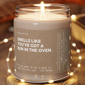 May include: A brown soy candle in a glass jar with a label that reads "Smells like you've got a bun in the oven". The label also includes the text "Hand-poured soy blend candle crafted in the USA, 50 hours burning time, 9oz glass jar".