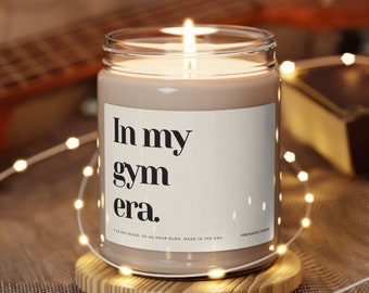 以下が含まれることがあります： 白いラベルに「In my gym era.」と書かれた茶色のキャンドル。キャンドルに火が灯され、木製のコースターの上に置かれています。キャンドルは白いストリングライトに囲まれています。
