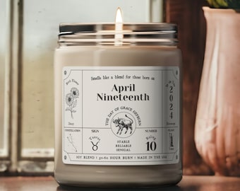May include: A brown glass candle jar with a white label. The label has the text "April Nineteenth" and a Taurus zodiac symbol. The candle is lit and has a white wax.